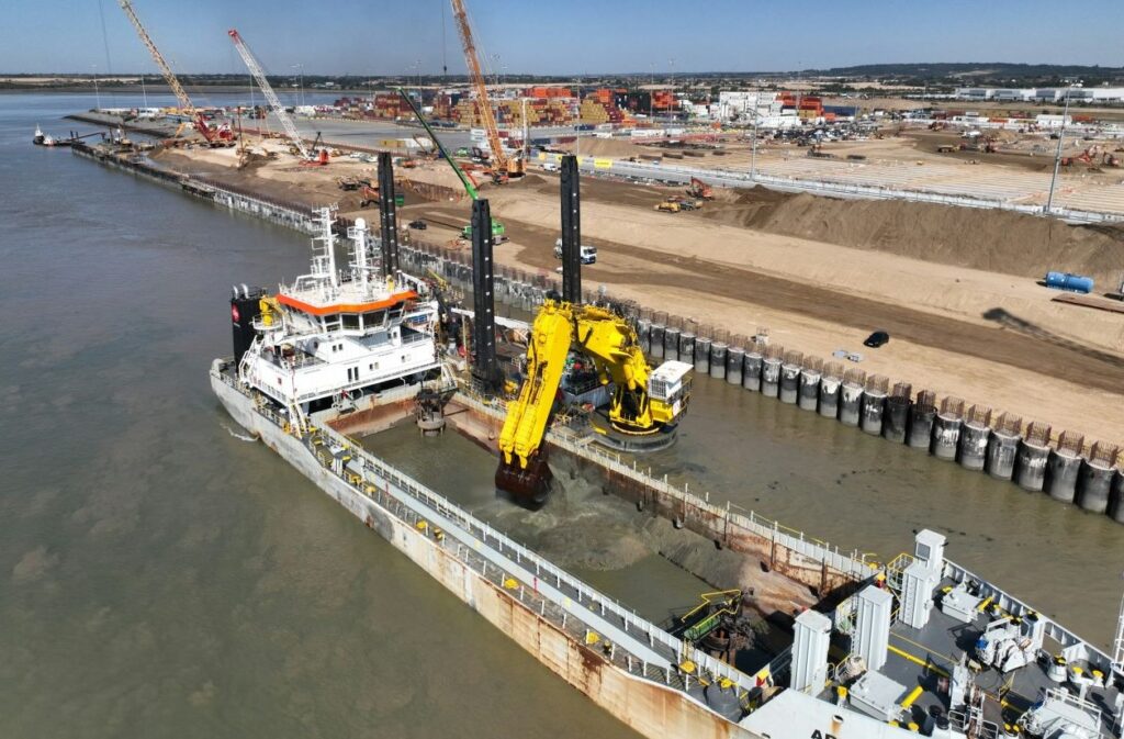 A large dredging vessel operating near a port, with cranes and containers visible in the background.